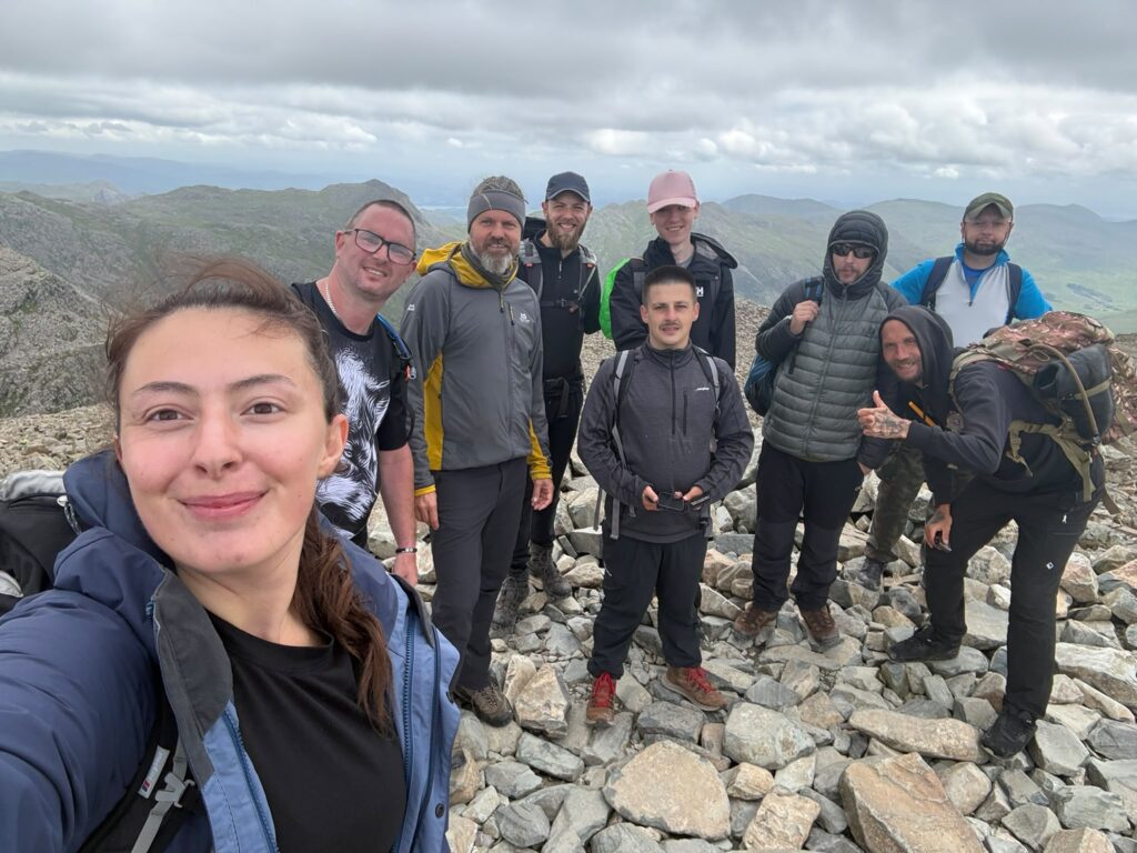 Grŵp o bobl yn gwenu mewn dillad awyr agored yn sefyll ar ben pentwr o gerrig, ar gopa Scafell Pike. Y tu ôl iddyn nhw, amrywiaeth o gopaon mynyddoedd ac awyr gymylog.