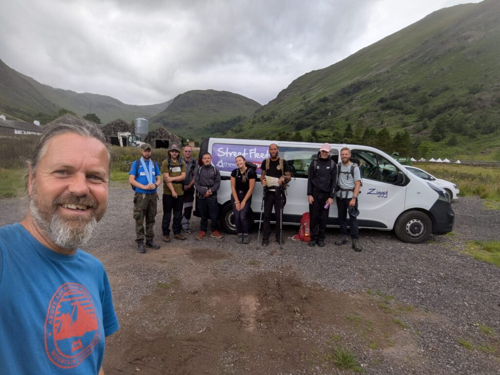 Grŵp o bobl mewn dillad awyr agored ac yn cario sachau teithio mewn rhes wrth ymyl fan fach gyda’r enw Ziggy wedi'i ysgrifennu ar yr ochr mewn porffor. Yn y cefndir mae bryn gwyrdd a bryn cymylog llwyd. Yn y pen blaen mae dyn â barf lwyd a chrys-t glas, yn gwenu ar y camera.