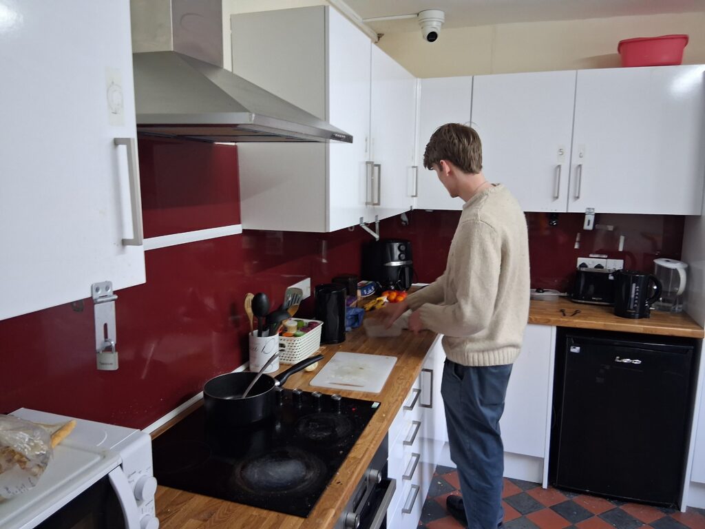 A young person wearing a beige jumper is cooking in a kitchen with white cupboards and a burgandy splashback