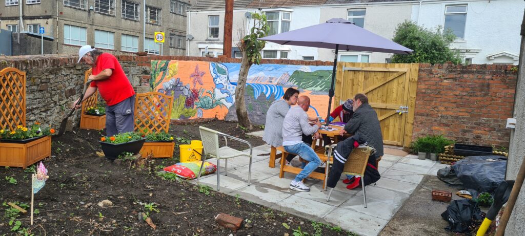Art session at The Wallich Shoreline Swansea. Four people are sitting around a table in a garden, under a parasol. One man in a red t-shirt is working in the garden.