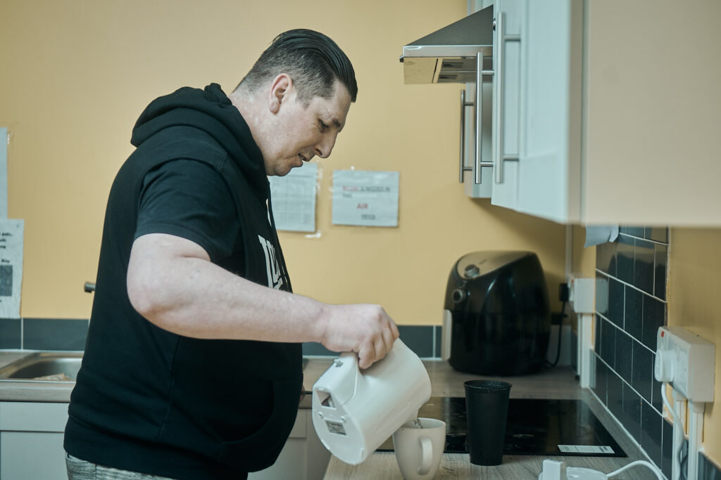 Man wearing a black hoodie pours water from a kettle into a mug. Behind him are kitchen units and appliances.