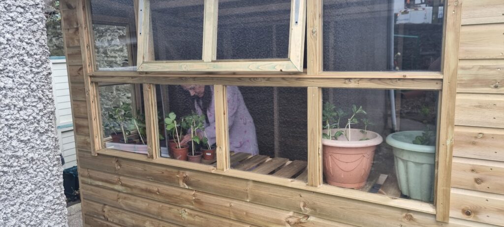 The Wallich Swansea Shoreline Garden project. A large wooden shed, through the windows are plant pots and a person looking after some seedlings.
