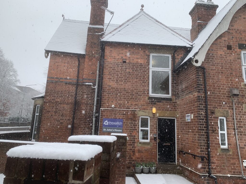 A red brick Victorian style building, with the roof and gate posts covered in snow. Snow is falling. A sign by the door reads St John's House. St John's Hostel