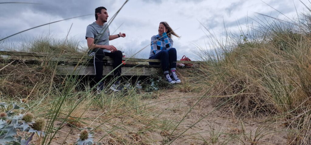 A woman with long hair talks with a dark-haired man wearing a grey t-shirt. They are sitting on a wooden walkway surrounded by beach plants. Drug & Alcohol Wellbeing Service (DAWS) Wrexham & Flintshire