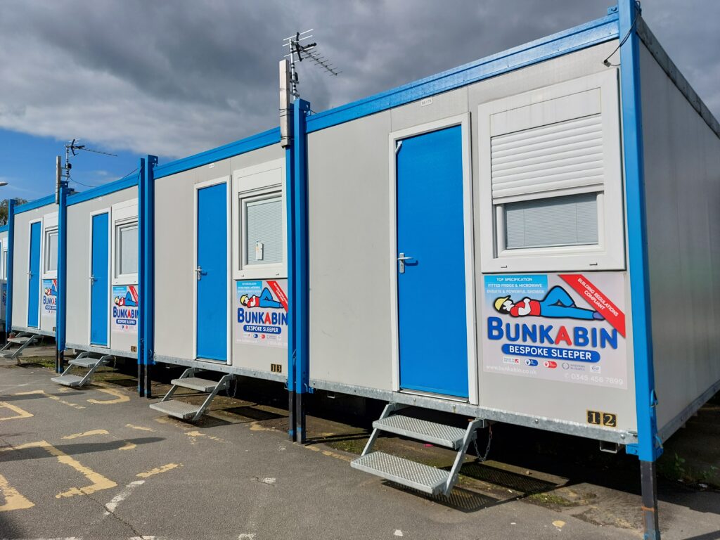 A row of four Bunkabin sleeping cabins, with metal steps up to a blue door and a window with a shutter.