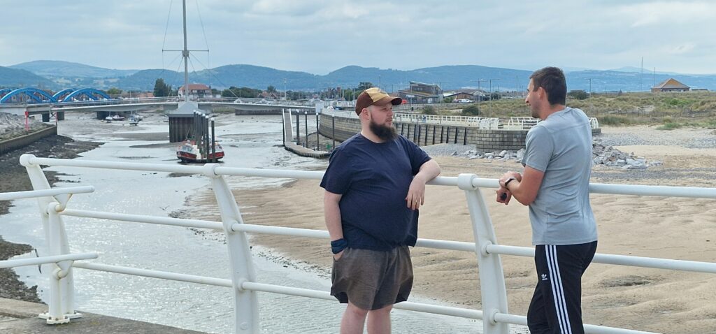 Two men chatting, leaning on white railings with sand, sea and a harbour in the background