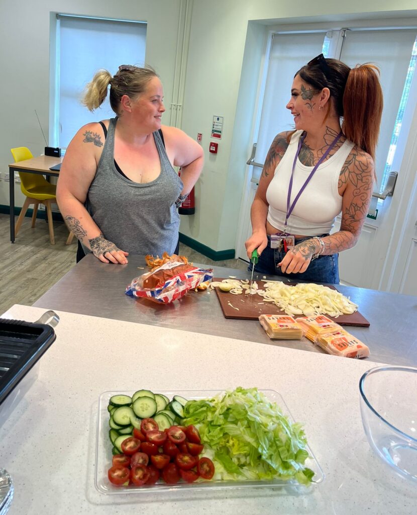 Two women, one blonde and one brunette, both wearing t-shirts, preparing food in a kitchen.