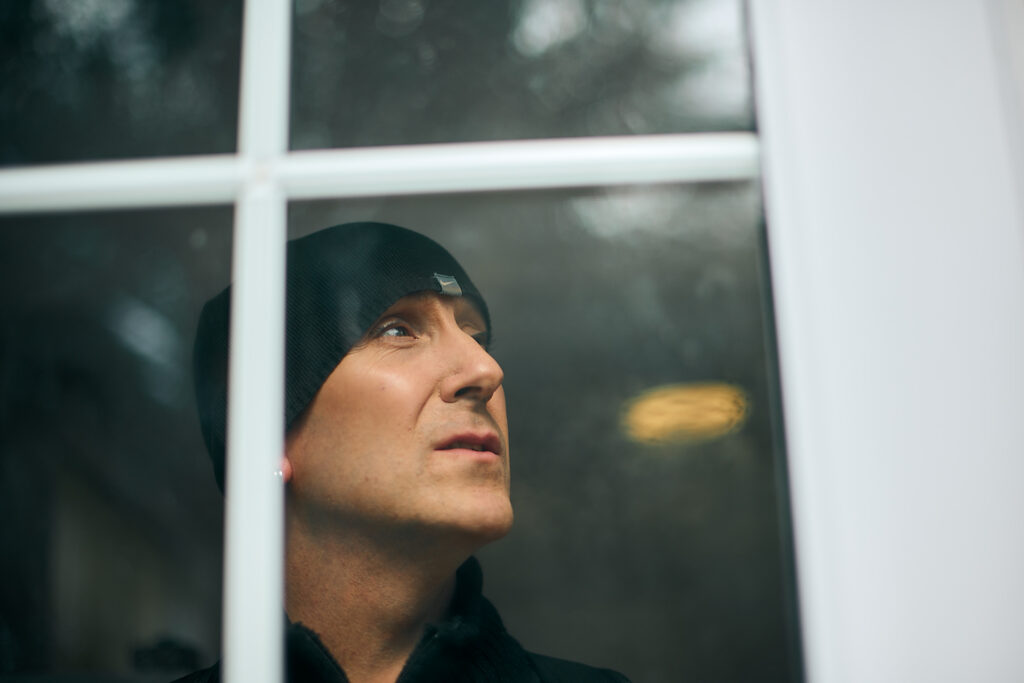 A man wearing a black beanie hat looks out through a window. His expression is thoughtful