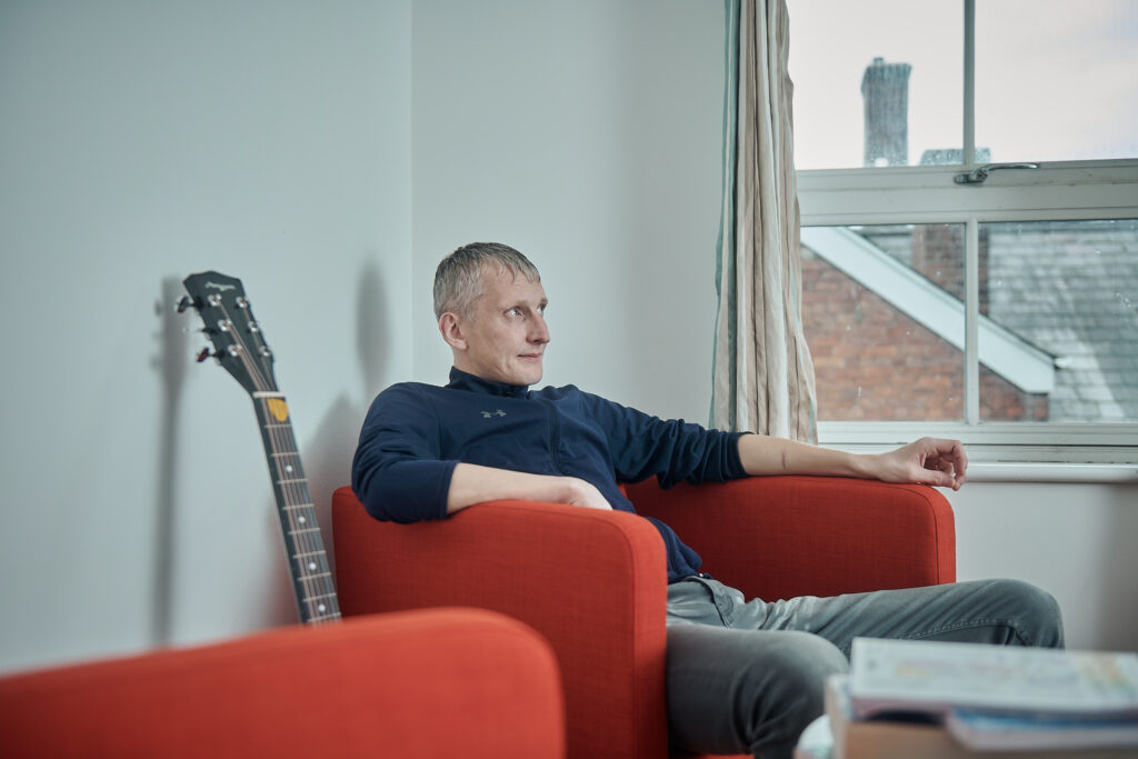A man with grey hair wearing a long-sleeved blue top sits in an orange arm chair. He is looking out of the window, with a guitar next to his seat. At Richmond House.