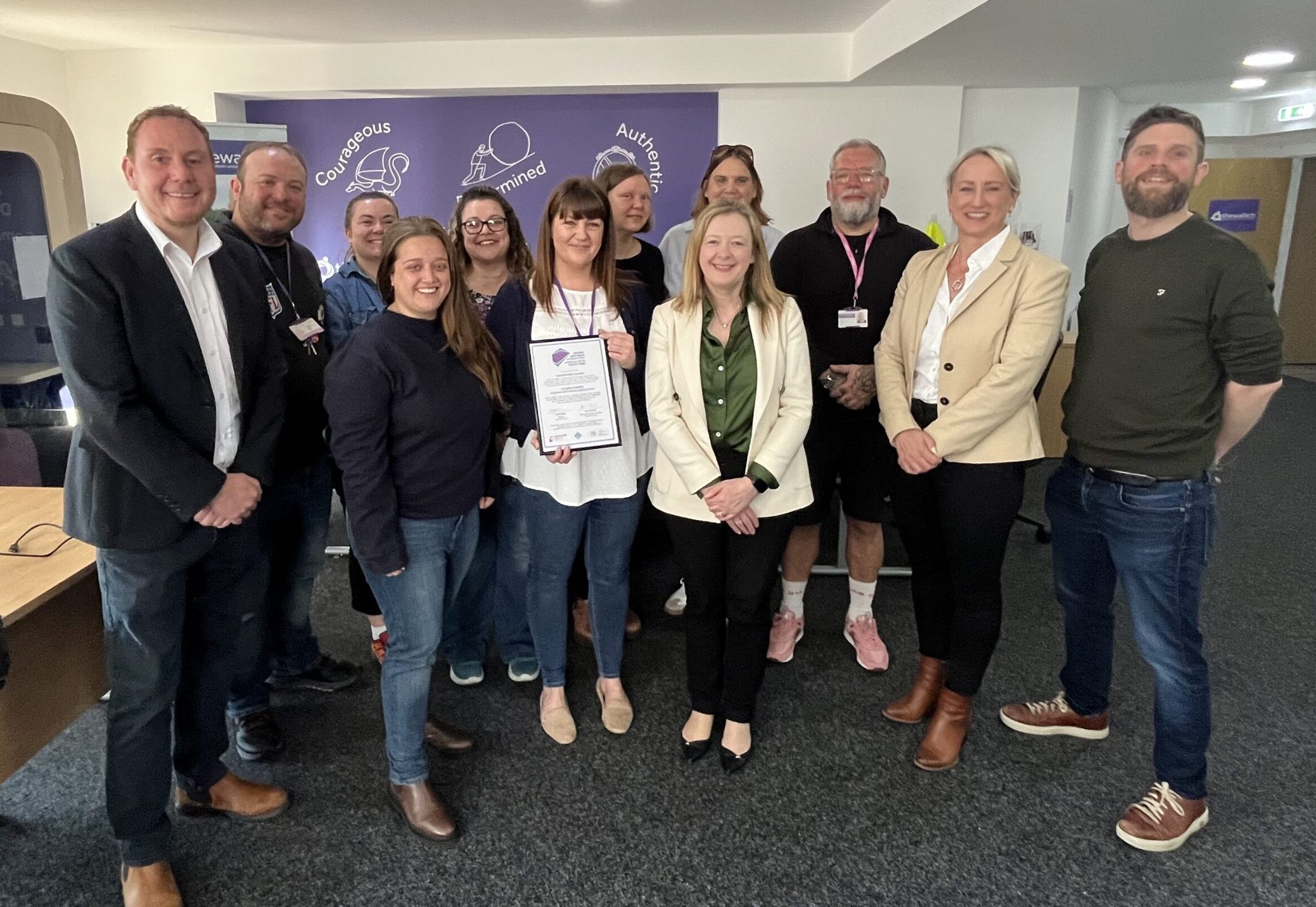 A group of people in an office gathered around a woman holding The Wallich Housing First Swansea Wales accreditation certificate
