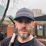 Volunteer man with a beard wearing a cap, with a garden tool over his shoulder