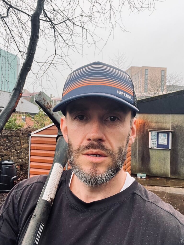 Volunteer man with a beard wearing a cap, with a garden tool over his shoulder