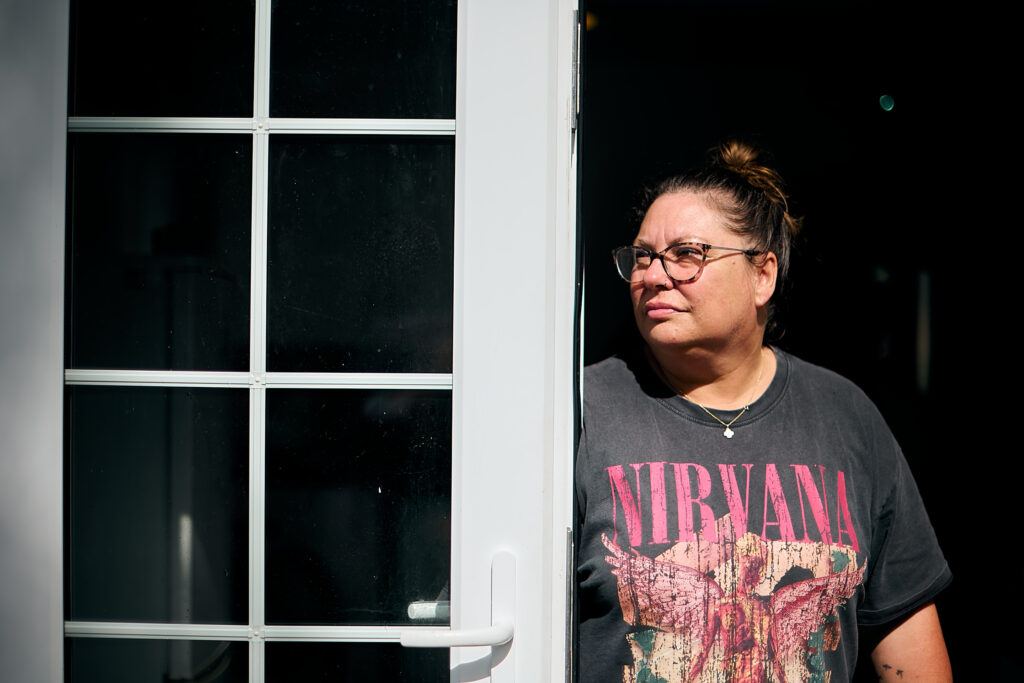 A woman wearing a black Nirvana t-shirt stands looking out of an open door. Her expression is calm and positive. 