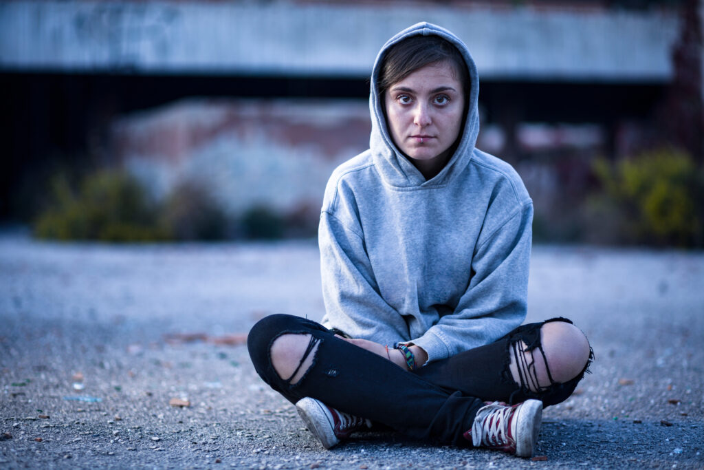 A young person with dark hair sits on the ground. They are wearing a grey hoodie, ripped black jeans and trainers. They are looking directly at the camera. Ketamine use is rising among young people in North Wales.