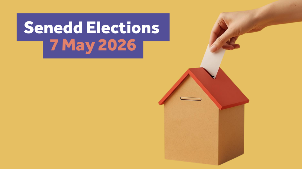 A hand holding a piece of paper is putting a vote into a ballot box shaped like a house