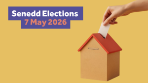 A hand holding a piece of paper is putting a vote into a ballot box shaped like a house