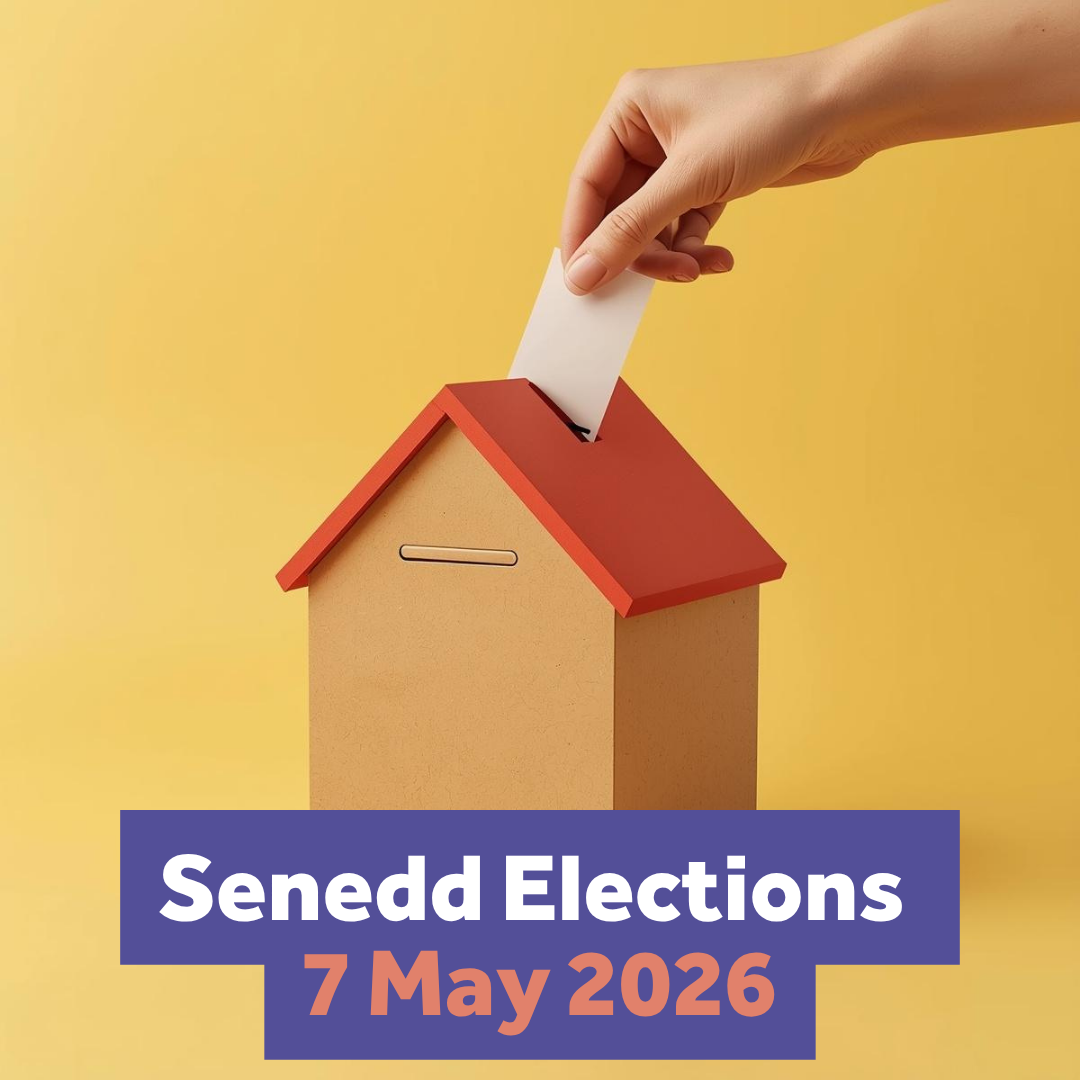 A hand holding a piece of paper is putting a vote into a ballot box shaped like a house