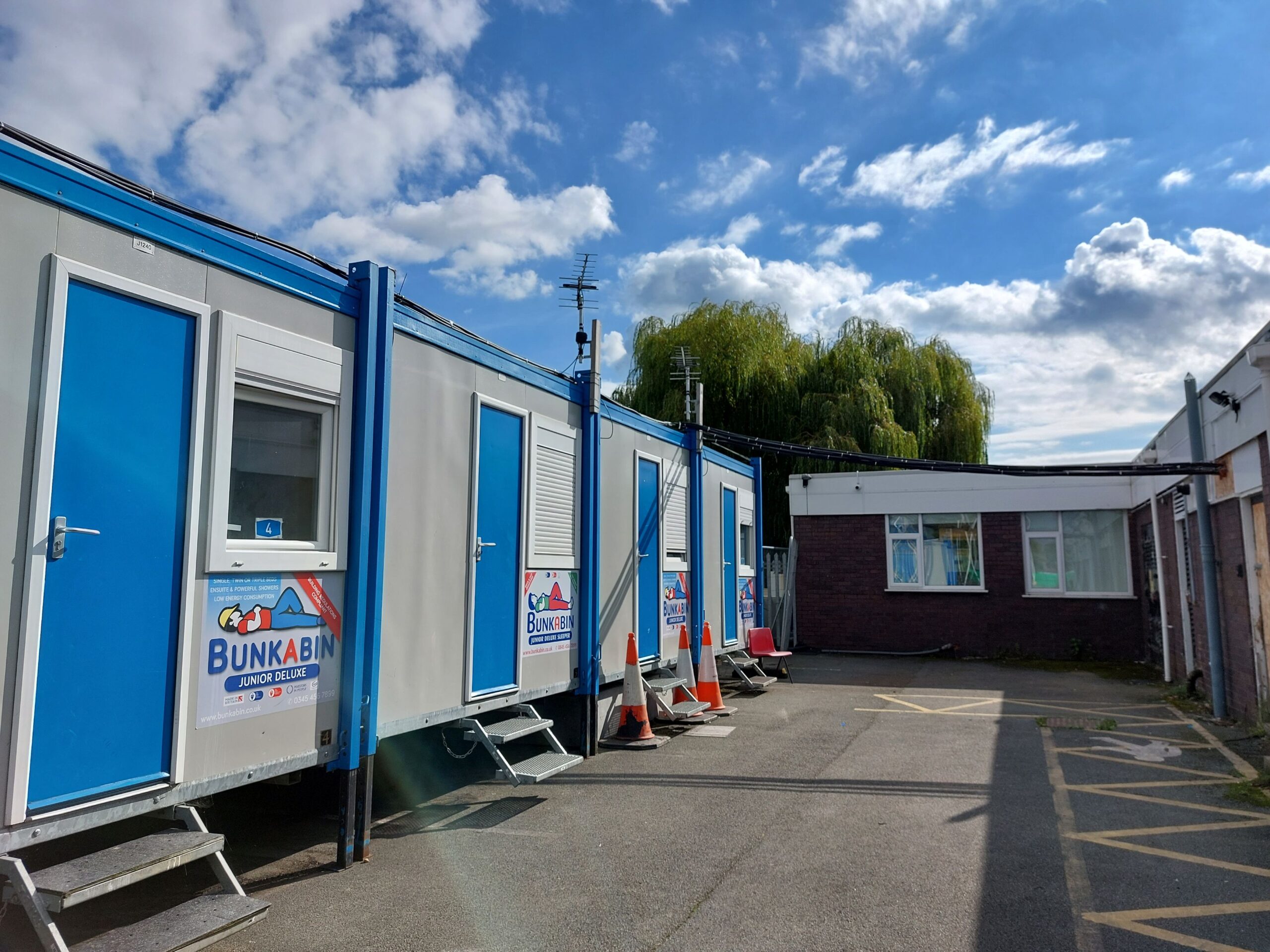 A row of Bunkabin cabins in grey with blue doors. In the background is a single story brick building. Glanrafon, Queensferry, Flintshire
