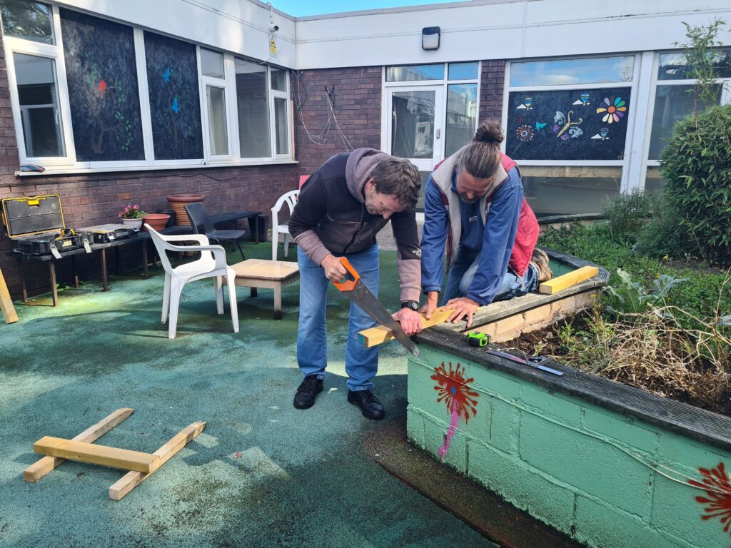 Two men doing woodwork in the garden of Glanrafon, Queensferry, Flintshire. They are cutting wood on a low wall, with a brick building behind them.