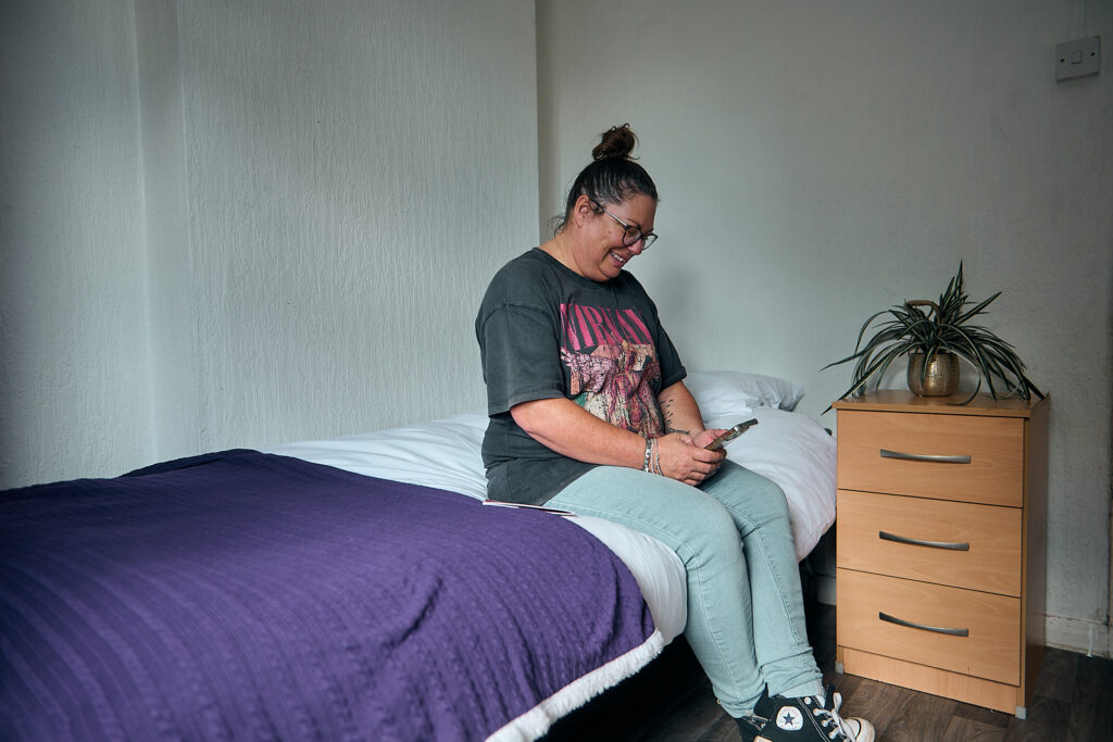Woman wearing grey band T-shirt and blue jeans, sitting on a bed looking at her phone. Temporary accommodation