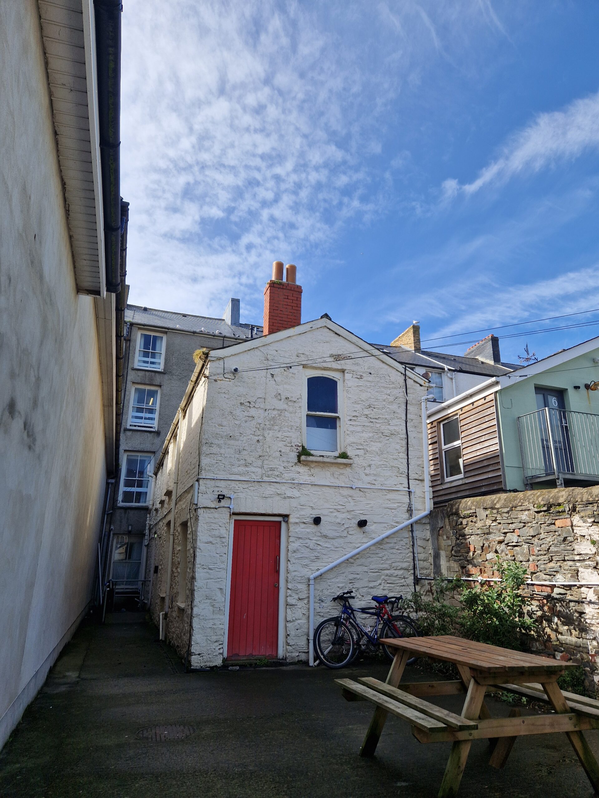 The back of a white-painted building with a red door. A bike and a bench are in front. Aberglasney