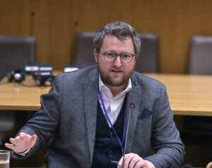 A man wearing glasses, a grey jacket and blue waistcoat sits behind a microphone. One hand is gesturing as he speaks.